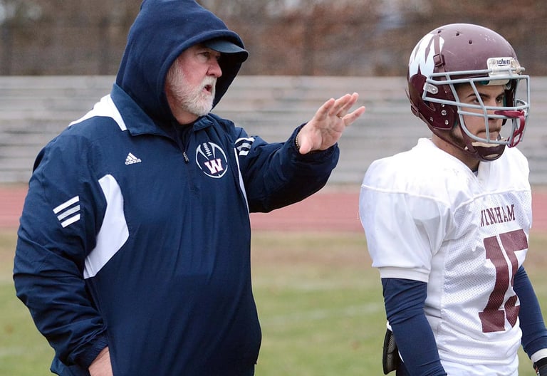 Football coach in navy Adidas jacket gesturing to player in white uniform on field
