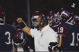 Football coach in white shirt holding up helmet with players in maroon uniforms standing behind him