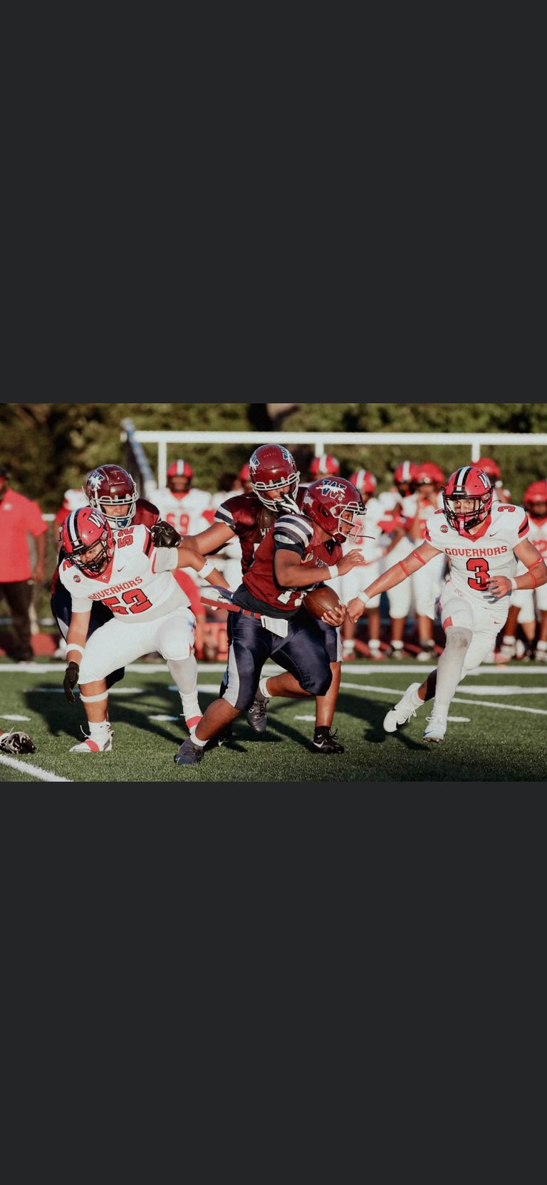 Young football player in maroon uniform running with ball while red and white team defenders close in during youth football game