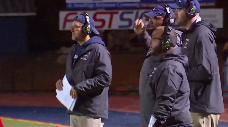 Football coaching staff gathered on sideline wearing team jackets and headsets during game