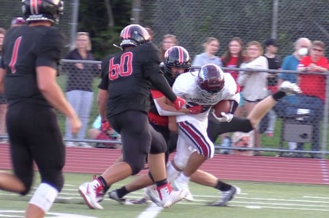 High school football player in maroon uniform evading black-uniformed defenders during game with spectators watching