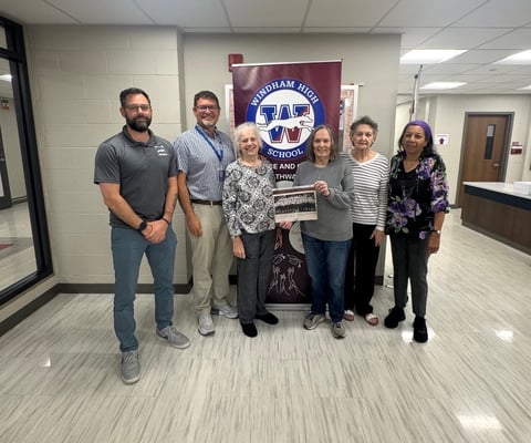 Seven people standing in a line indoors in front of a Windham High School banner, one person holding a plaque