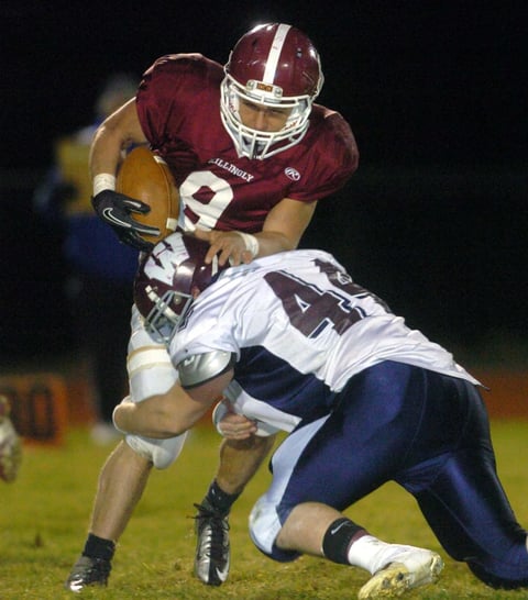 Football player in maroon jersey carrying ball while being tackled by defender in blue and white uniform during night game