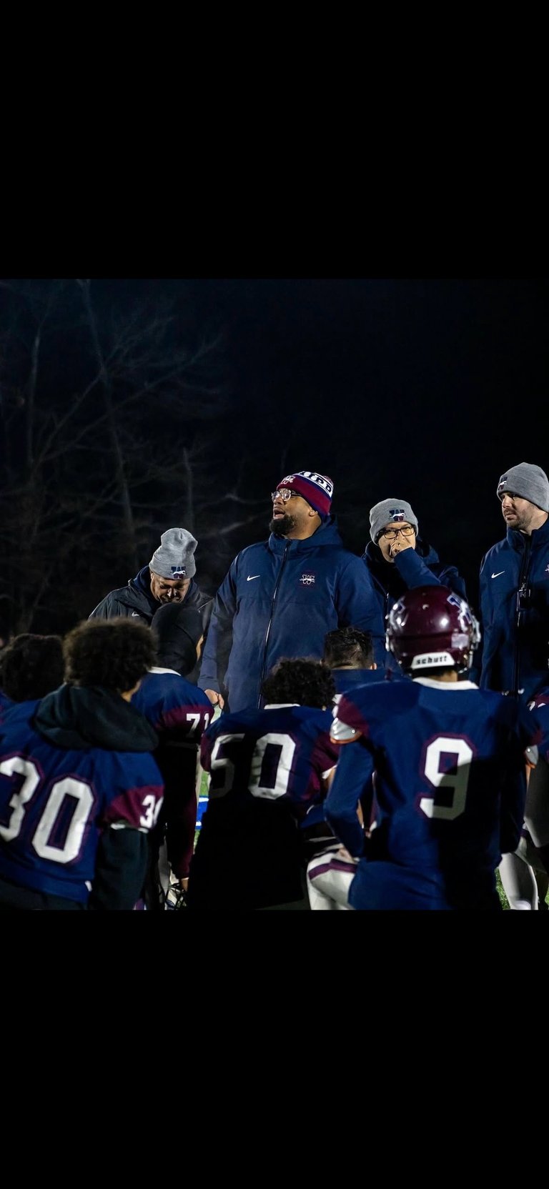 Football coaches in blue winter gear huddle with players wearing navy and maroon uniforms during a nighttime practice or game.