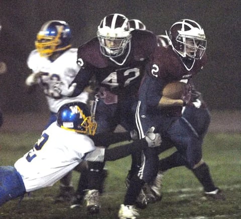 Football players in blue and white uniforms tackling a player in navy jersey number 42 during a night game