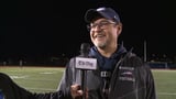 Man holding microphone on football field wearing glasses, cap, and Windham High School Football jacket
